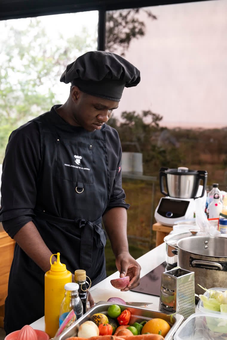 A contestant in a black chef’s jacket chops onion at his work table.