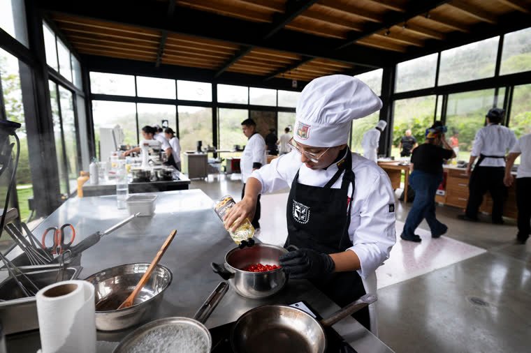 Wide view of the kitchen with a contestant adding a liquid to a pot.