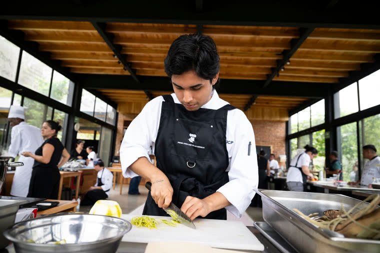 A young chef cuts ingredients at his station, under the hall’s wooden roof.