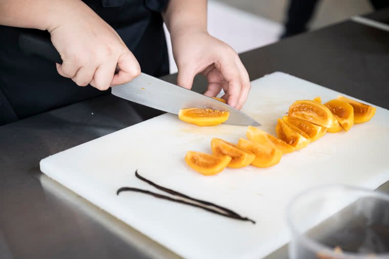 A knife cutting wedges of yellow fruit beside two vanilla pods.