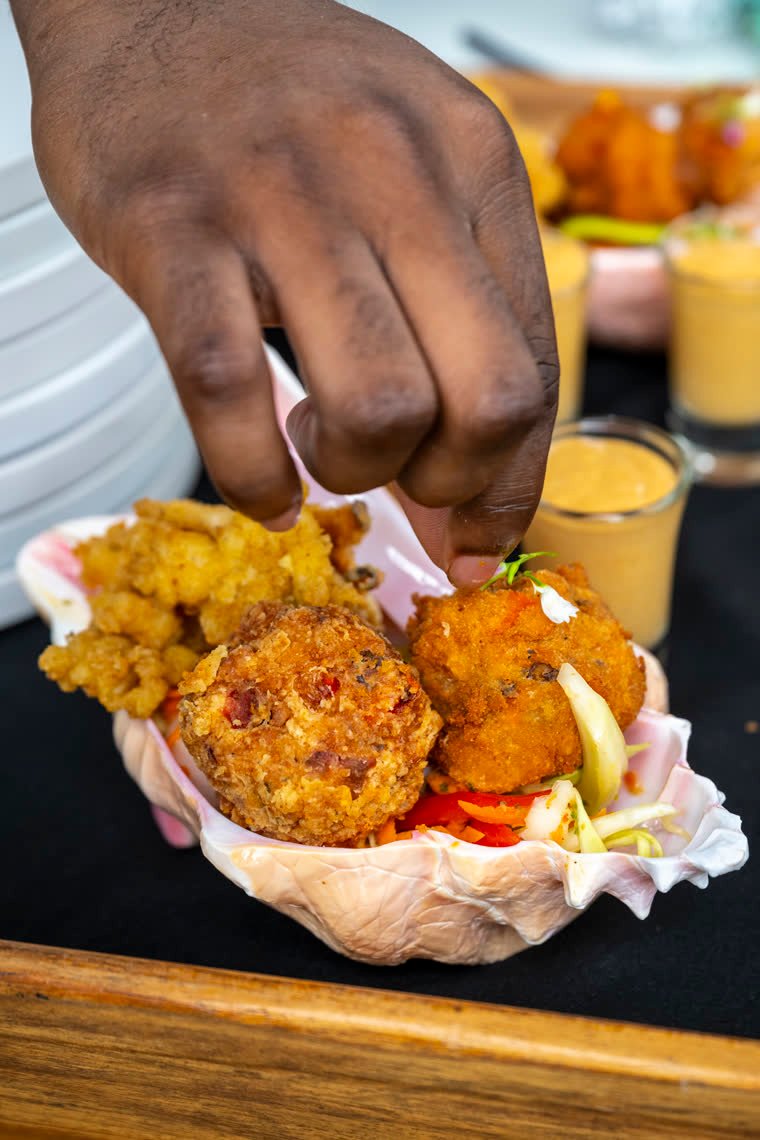 A hand adjusts the finish of croquettes served in a shell, with sauces in the background.