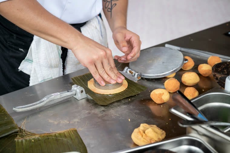 Hands shape dough in a press over a banana leaf; golden pieces beside it.