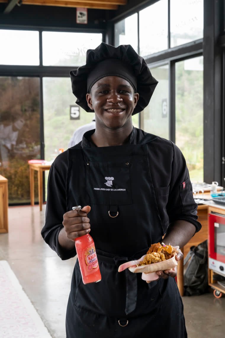 A contestant smiles holding his dish in a shell and a bottle of a drink.