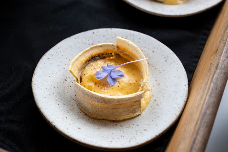 A small golden tartlet with a violet flower on a speckled ceramic plate.