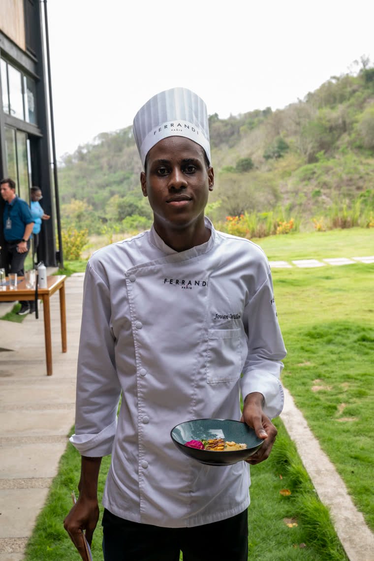 A chef in a tall hat holds his dish in a garden.