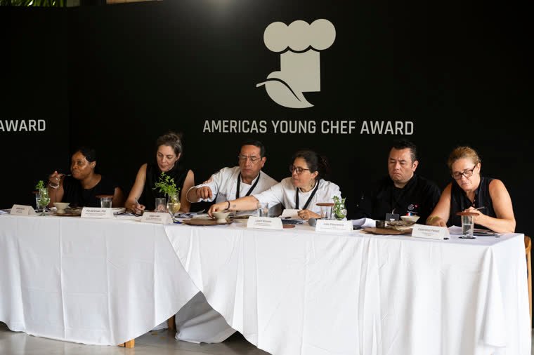Six judges evaluate the dishes in front of the Americas Young Chef Award wall.