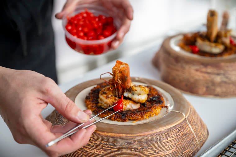 Plating detail with tweezers and a small red fruit on a shellfish dish.