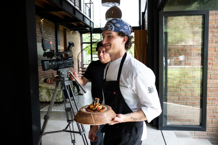 The contestant carries his finished dish; a video camera in the background.