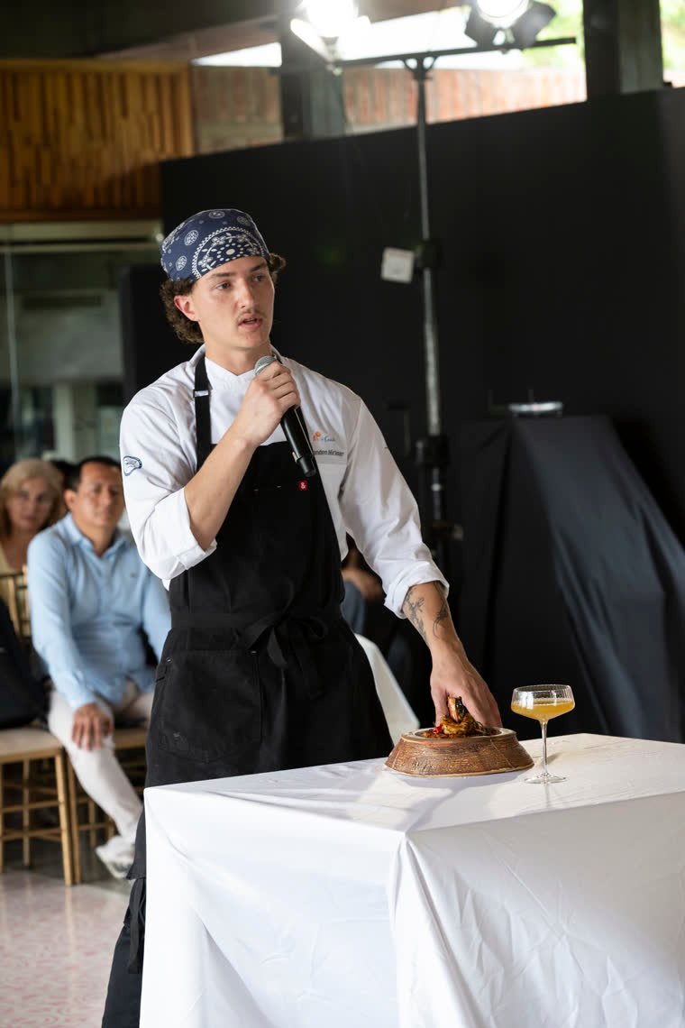 A contestant in a blue bandana presents his dish on the table, next to a cocktail.