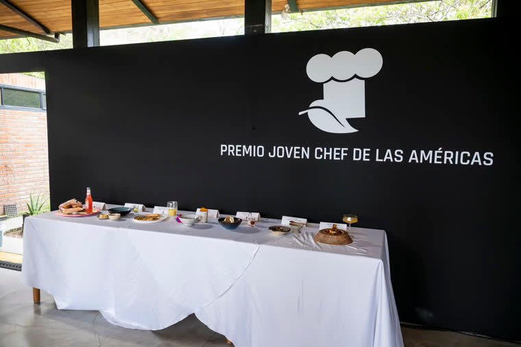Table with the competition dishes in front of the Americas Young Chef Award wall.